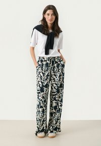 White short-sleeve shirt with a navy sweater tied at the neck, paired with loose blue and cream patterned trousers. Casual footwear visible.