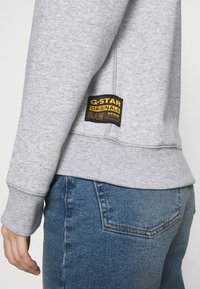 Felpa grigia in morbido tessuto, con polsini a coste. Etichetta G-Star nera e gialla cucita nella parte bassa della schiena, abbinata a jeans blu.