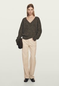 Kobieta z prostymi włosami, ubrana w ciemny sweter z dekoltem w serek, beżowe spodnie, czarne buty, trzymająca ciemną kurtkę w jednej ręce, stojąca na jednolitym tle.