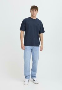 Camiseta de algodón azul marino con mangas cortas y cuello redondo, que presenta un pequeño logotipo bordado en el pecho, combinada con unos vaqueros de color azul claro y zapatillas blancas.