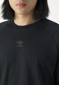 Black cotton long-sleeve shirt with a round neckline and raglan sleeves, featuring a subtle black logo on the chest. Soft texture, regular fit.