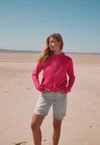 Sweat à capuche zippé rose avec une finition texturée, associé à un short en jean bleu clair déchiré, sur fond de plage sablonneuse.