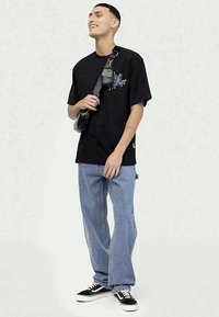 Black cotton t-shirt with blue graphic, paired with light blue denim jeans and black sneakers. A crossbody bag hangs from the shoulder.