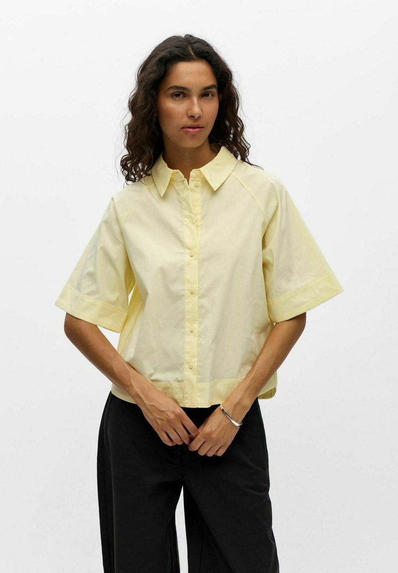 Femme aux cheveux bouclés portant une chemise jaune clair à manches courtes boutonnée et un pantalon noir, debout les mains jointes devant elle.