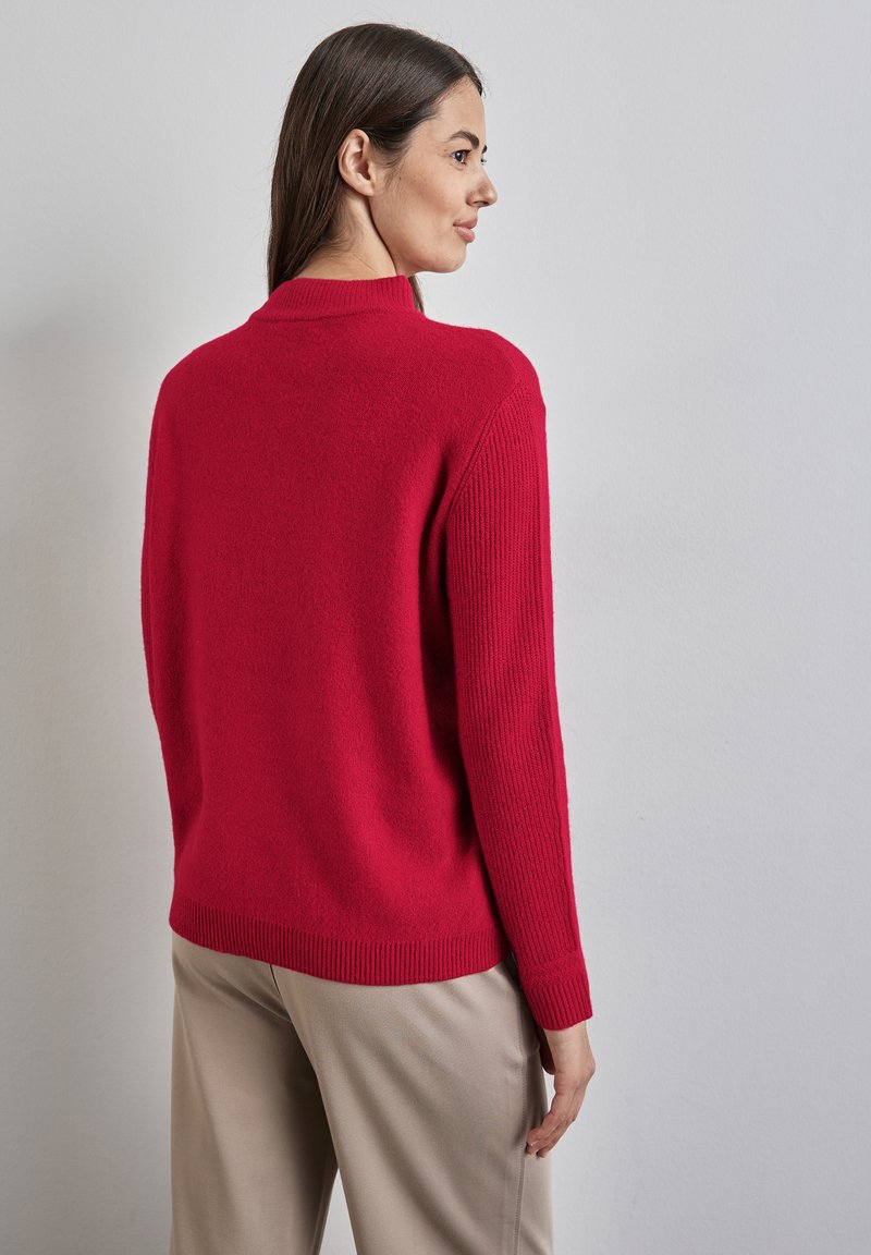 Maglione rosso lavorato a maglia con polsini e orlo a coste. Presenta un collo alto ed è abbinato a pantaloni beige. Texture morbida visibile.