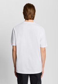 Άτομο με μεσαίου μήκους καστανά μαλλιά φοράει απλό λευκό κοντομάνικο πουκάμισο και μαύρο παντελόνι, με την πλάτη γυρισμένη προς ένα απλό γκρι φόντο.