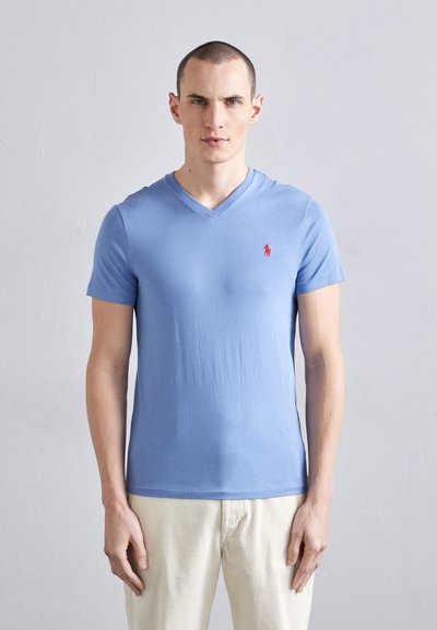 Sininen lyhythihainen V-neck T-paita, joka on valmistettu pehmeästä kankaasta ja jossa on pieni punainen polo-logo vasemmassa rinnassa, yhdistettynä vaaleisiin housuihin.