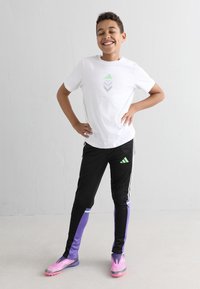 Hvid t-shirt med grønne og lilla geometriske mønstre, parret med sorte træningsbukser med lilla sideaccenter og lyserøde løbesko.