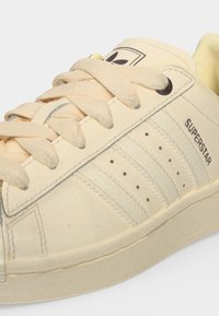 Κρεμ χρωματιστό παπούτσι Adidas Superstar με υφασμένα κορδόνια, τρεις ραμμένες ρίγες και το κείμενο "SUPERSTAR" στο πλάι.