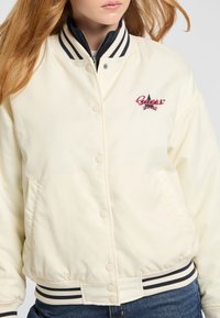 Crème bomberjack met een geribde kraag, drukknoopsluiting en geborduurd logo in rood en zwart. Voorzien van gestreepte manchetten en een tailleband.