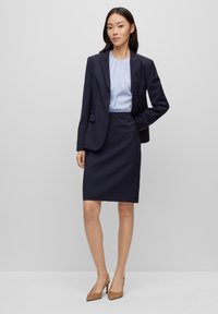 Costume marine avec un blazer ajusté, blouse bleu clair et jupe crayon ; tissu lisse, coupe ajustée, associé à des chaussures à talons hauts beiges.
