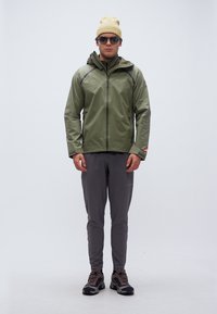 Άνδρας που φοράει γιλέκο με κουκούλα ελιάς, γκρι παντελόνι, παπούτσια πεζοπορίας, μπεζ σκούφο και γυαλιά ηλίου, στέκεται μπροστά σε απλό φόντο.