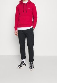 Röd bomullshuvtröja med snoddar och framficka, lager på en vit skjorta. Kommer med svarta byxor och svarta sneakers.