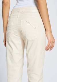 Pantalones beige claro con un ajuste slim, que presentan dos bolsillos traseros diagonales y detalles de costura sutiles. Tejido suave y texturizado con un acabado limpio.