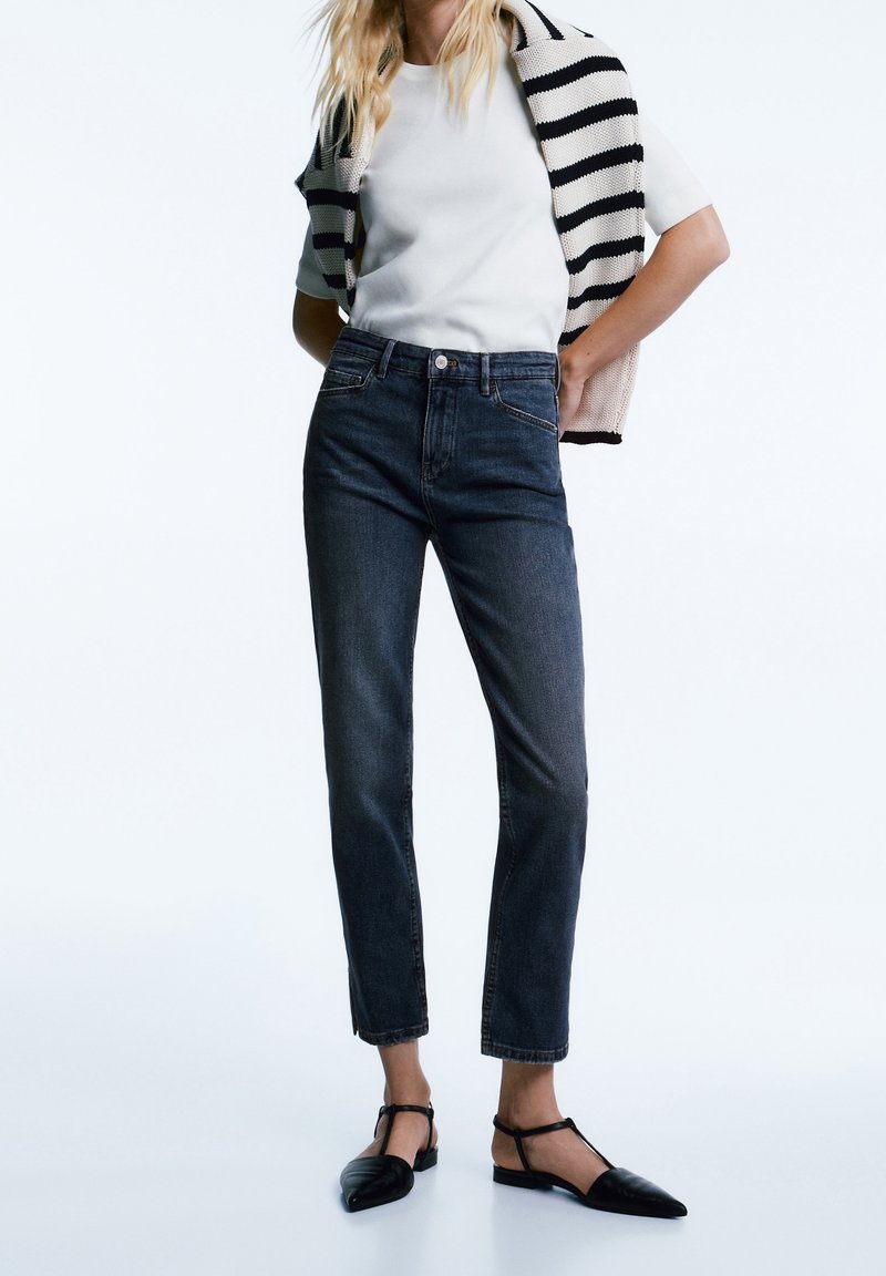 Jean taille haute en denim foncé avec une coupe droite, associé à un t-shirt blanc et à un cardigan en maille rayé noir et blanc. Chaussures plates noires.