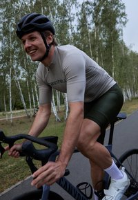 Cycliste portant un maillot beige ajusté avec un short vert, roulant sur un vélo gris. Il porte un casque noir et des guidons noirs sur un chemin pavé.