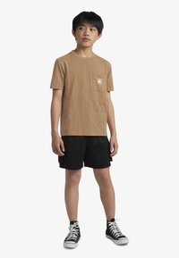 Rjava bombažna majica s hlačnim žepom in belim logotipom, kombinirana s črnimi mrežastimi kratkimi hlačami in črnimi visokimi supergami. Preprost dizajn, priložnostna prileganje.