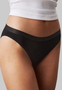 Crne Calvin Klein tange napravljene od glatkog tkanja, s širokim pojasom na kojem je suptilno utisnut naziv brenda.