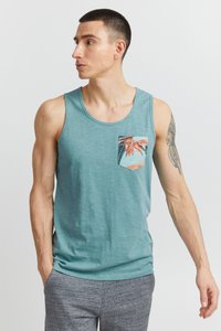 Tyrkysové tielko z ľahkej látky s predným vreckom s farebnou tropickou potlačou. Pod ním sú nosené sivé šortky.