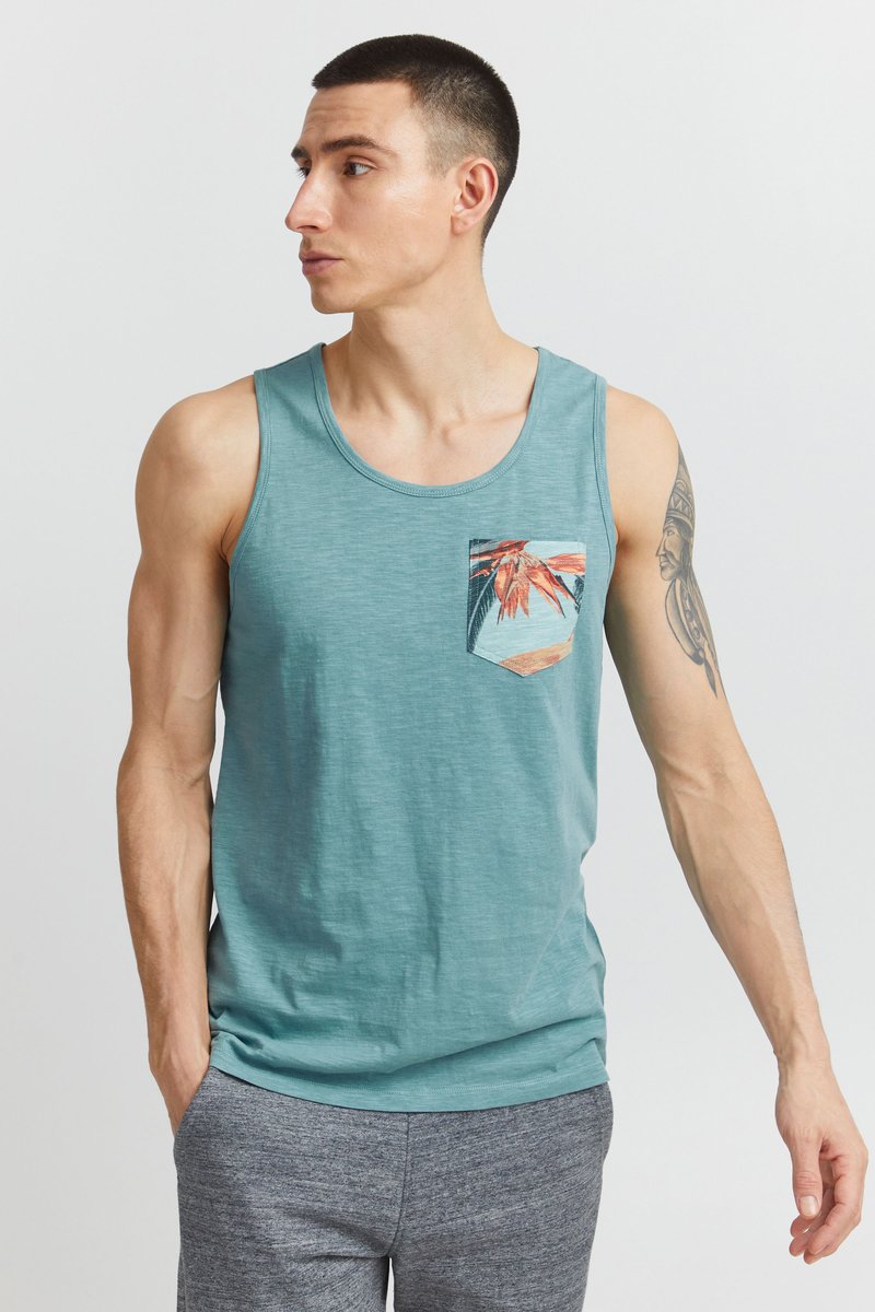 Tyrkysové tielko z ľahkej látky s predným vreckom s farebnou tropickou potlačou. Pod ním sú nosené sivé šortky.