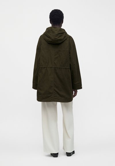 Personne aux cheveux courts et bouclés portant une veste à capuche vert olive et un pantalon blanc à jambes larges, debout dos à un fond uni.