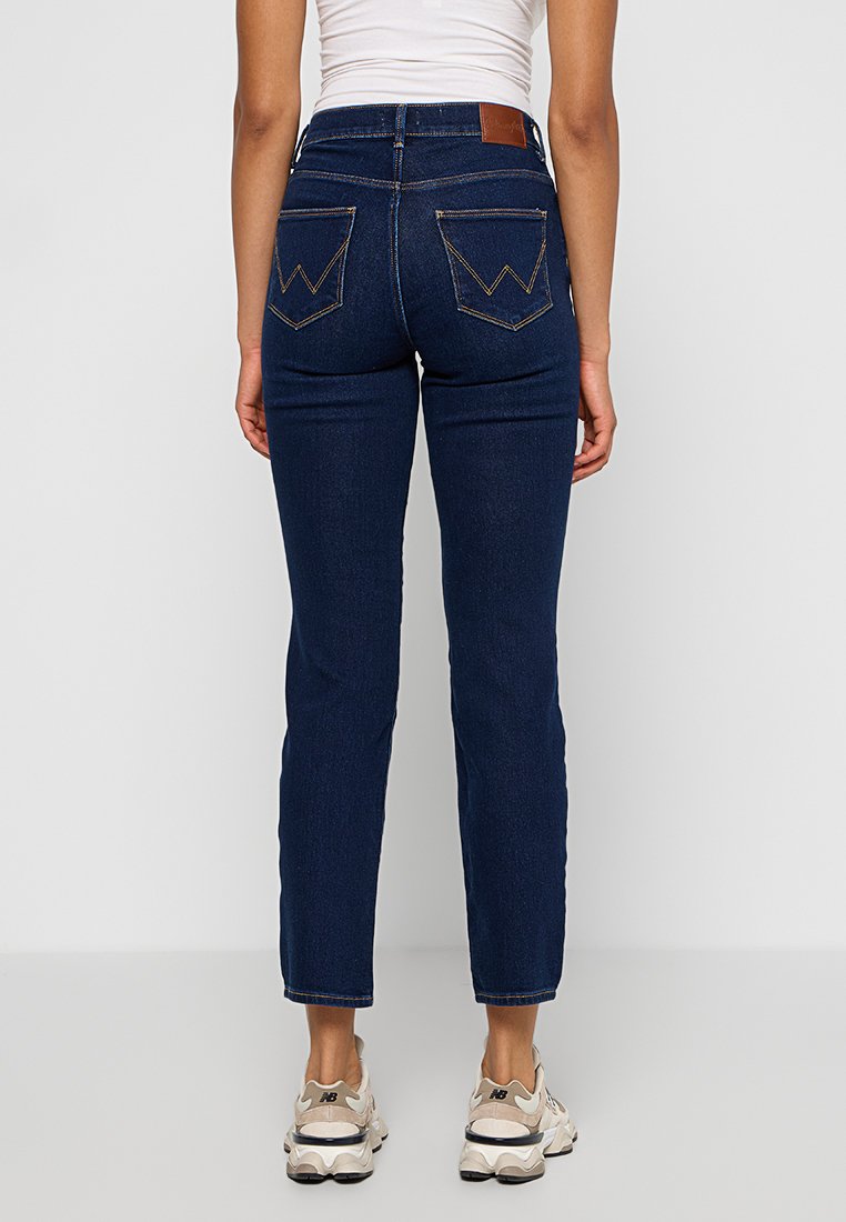Jean en denim bleu foncé à taille haute, coupe droite et coutures contrastantes. Dispose de poches arrière avec un détail en zigzag.