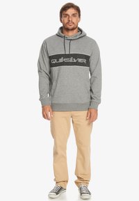 Grijze hoodie met zwarte streep en "Quiksilver" logo in witte letters. Draag het met beige broek en zwarte sneakers.