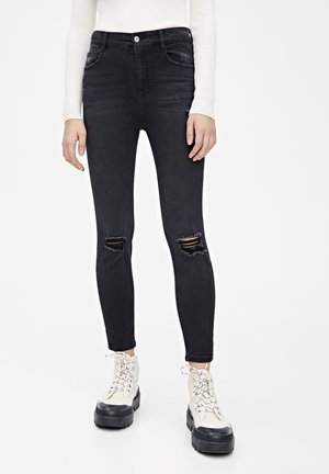 Kvinde iført sorte slidte skinny jeans med revne knæ og hvide højtop-platformstøvler, stående mod en hvid baggrund.