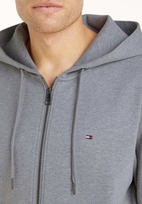 Felpa con cappuccio grigia con cerniera, realizzata in tessuto morbido, dotata di cappuccio con coulisse, cerniera frontale e piccolo logo sull'area sinistra del petto.