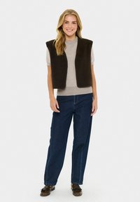 Mujer sonriente con pelo largo y rubio, vestida con una camiseta corta color beige, un chaleco sin mangas marrón oscuro, jeans azules y mocasines marrones, de pie.