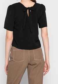 Top nero a trama con maniche a palloncino e retro con apertura a chiave, legato con un fiocco. Abbinato a pantaloni marroni con tasche posteriori e cuciture a contrasto.