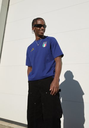 Blauw sport T-shirt met het Italië-embleem en gouden accenten, gecombineerd met zwarte corduroy cargobroek. Model draagt een zonnebril en staat buiten.
