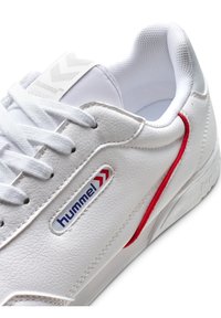 Fehér sneaker, textúrált bőr külsejével, piros díszcsíkkal, fehér fűzőkkel és egy logóval a nyelv közelében.