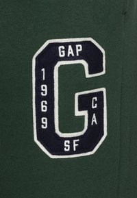 Tmavě zelená mikina s vyšitým námořnickým modrým znakem s písmenem "G", rokem "1969" a textem "GAP CA SF" v bílé barvě.