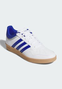 Zapatillas deportivas blancas de cuero sintético con detalles azules y tres rayas. Presentan una suela de goma marrón y una lengüeta acolchada azul.