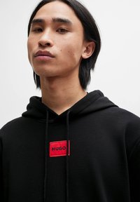 Svart hettegenser laget av bomull, med en rød rektangulær lapp med "HUGO" tekst. Glatt tekstur, snøring ved halsen.