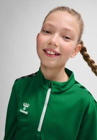 Groene, textuurachtige sportjack met hoge kraag, witte rits en zwarte accenten op de schouders. Logo aan de linkerkant weergegeven.