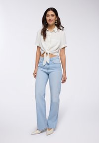 Camicia bianca a maniche corte, con colletto e bottoni, con un nodo frontale; jeans a vita alta azzurri chiari; tacchi beige appuntiti. Tessuto dalla superficie liscia.