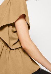 Blusa leggera color beige con design drappeggiato, maniche corte e vita elastica. Texture liscia con delicate pieghe lungo la vita.