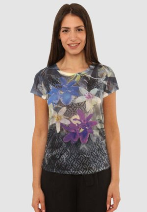 Giovane donna sorridente, indossa una camicia a maniche corte con motivi floreali blu, bianchi e viola su uno sfondo nero geometrico.