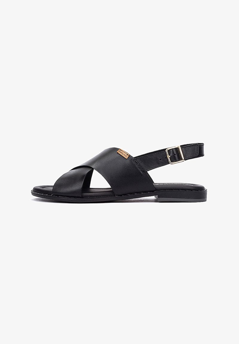 Sandale plate en cuir noir avec brides croisées, sangle de talon réglable avec boucle, et petite étiquette de marque sur le côté.