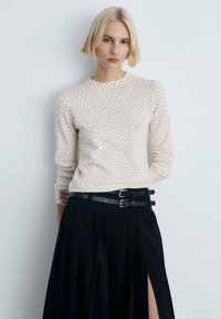 Cremefarbener Pullover mit schwarzen Punkten, Rundhalsausschnitt und langen Ärmeln. Kombiniert mit einem schwarzen gefältelten Rock mit hohem Schlitz und doppeltem Schnallen-Gürtel.