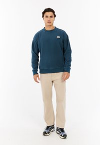 Diep teal sweatshirt met een ronde hals en geribbelde manchetten, gecombineerd met lichtbeige broek en zwart-blauwe sneakers met patroon.