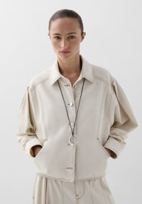 Beige oversized shirt jacket with button front, pointed collar, and loose sleeves; features contrast stitching and a pocket at the front.