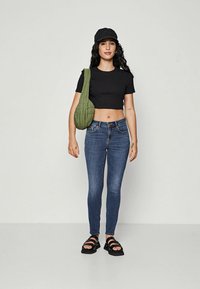 Black cropped t-shirt, blue skinny jeans, black sandals, green quilted shoulder bag, and a black cap. Smooth textures and fitted design.
