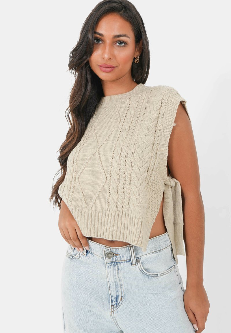 Pull collyre beige tricoté avec motifs en relief, emmanchures larges et ourlet côtelé, associé à un short en denim délavé.