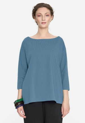 Kobieta ubrana w luźną, niebieską bluzkę z długim rękawem i czarne spodnie, trzyma na lewej ręce zielone, ciemnobrązowe i fioletowe bransoletki, stoi na białym tle.