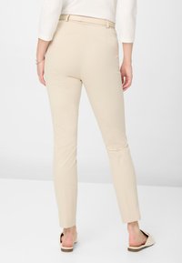 Pantalons beige ajustés avec une coupe slim. Texture lisse, taille mi-haute et un détail de couture subtil à la cheville. Portés avec des sandales claires.
