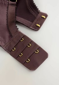 Primo piano di una chiusura di reggiseno in tessuto marrone con tre file di ganci e occhielli in metallo dorato su sfondo chiaro.