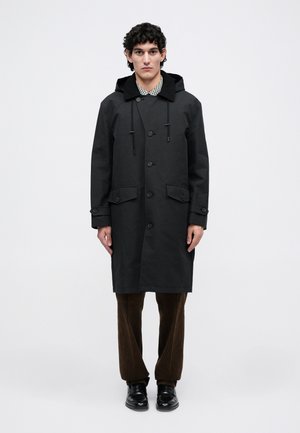 Man staat rechtop en kijkt naar voren, gekleed in een zwart knopenjas met kap, bruine corduroybroek en zwarte leren schoenen, op een witte achtergrond.